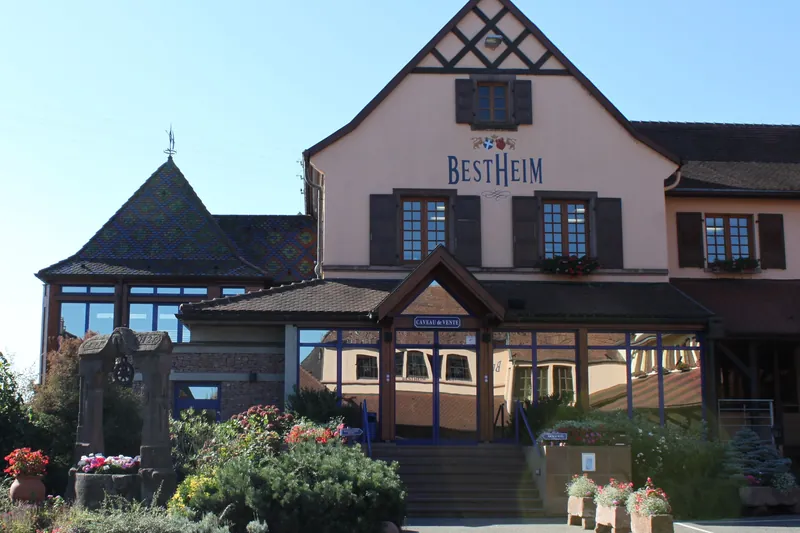 Bâtiment à colombages de la cave Bestheim à Bennwihr, avec son enseigne et l'entrée vitrée de la boutique, entouré de massifs fleuris