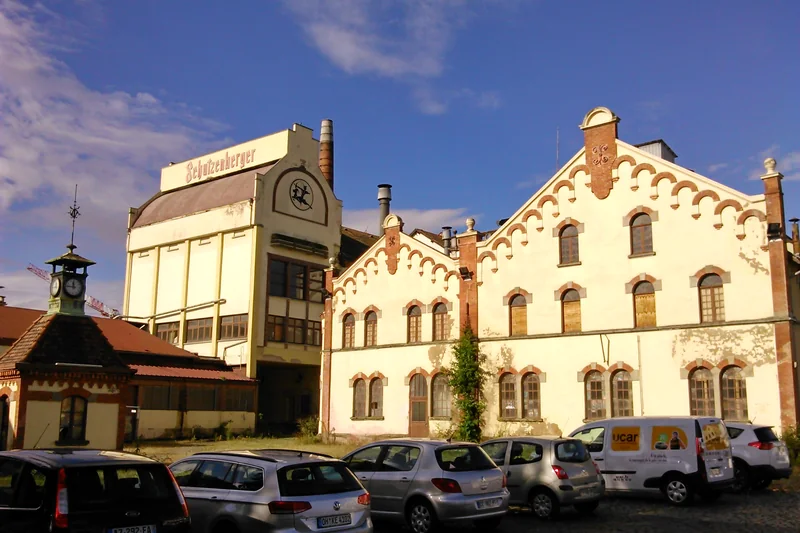 La brasserie Schutzenberger à Schiltigheim : bâtiments aux pignons ouvragés et grande enseigne, voitures garées devant.