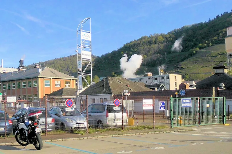 L'usine chimique de Thann et Vieux-Thann, avec ses bâtiments, sa tour et ses fumées blanches devant les vignes du coteau.