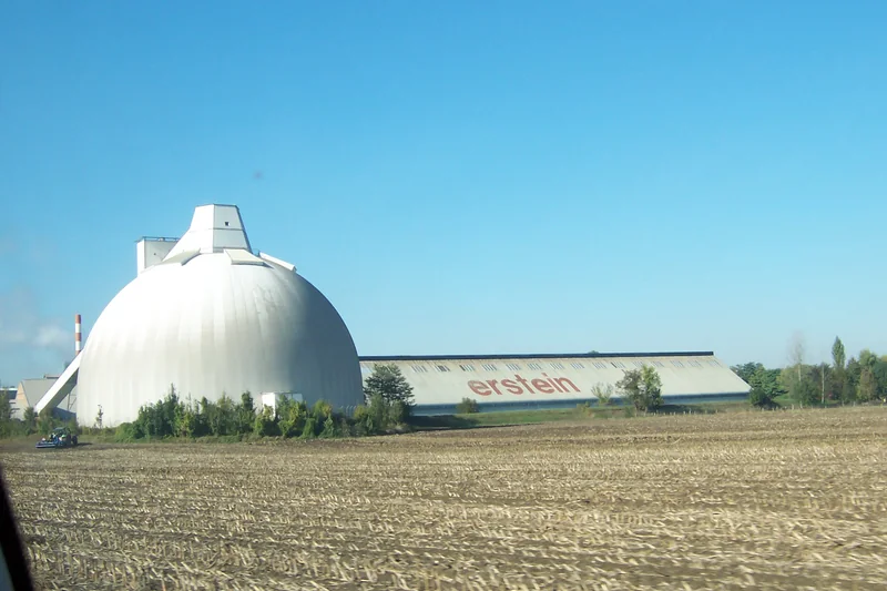 La sucrerie d'Erstein : un grand silo blanc en dôme et un long bâtiment portant le nom de la ville, derrière un champ.