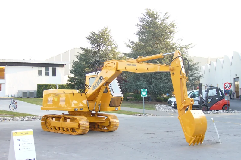 Une pelle hydraulique Liebherr jaune exposée dans la cour de l'usine de Colmar, avec des halls et un chariot élévateur derrière.