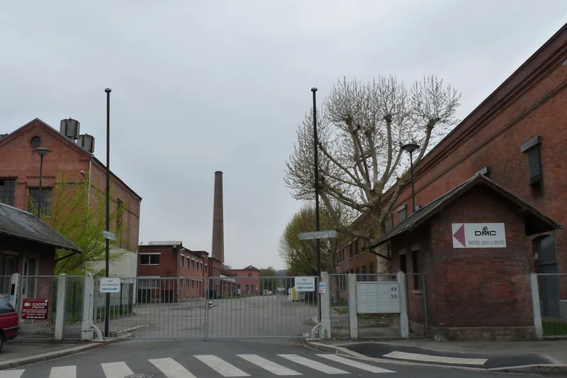 L'entrée du site DMC à Mulhouse : bâtiments de brique rouge et haute cheminée derrière la grille, en avril 2012.