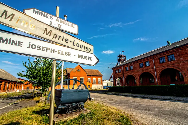 Panneaux indiquant les mines Amélie et Marie-Louise et le musée Joseph-Else, devant un chevalement et des halles de brique.